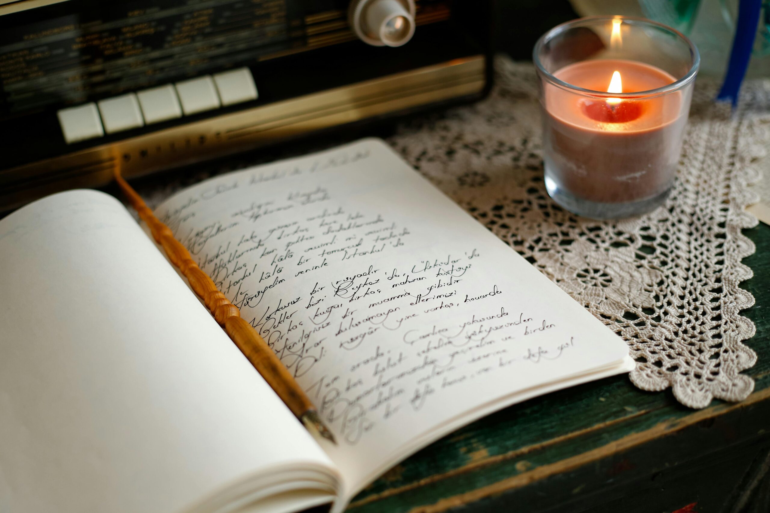 A warm, intimate scene showing a handwritten notebook, candle, and vintage radio on a lace tablecloth.