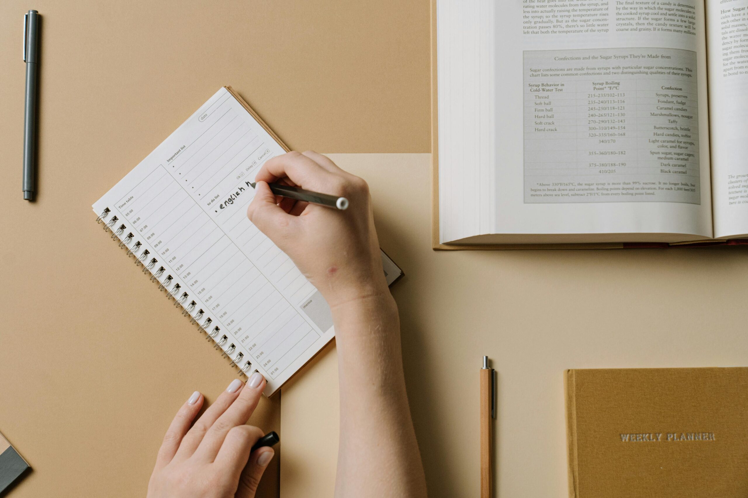 A person's hands writing in a planner at a desk with an open book nearby, conveying an organized workspace.
