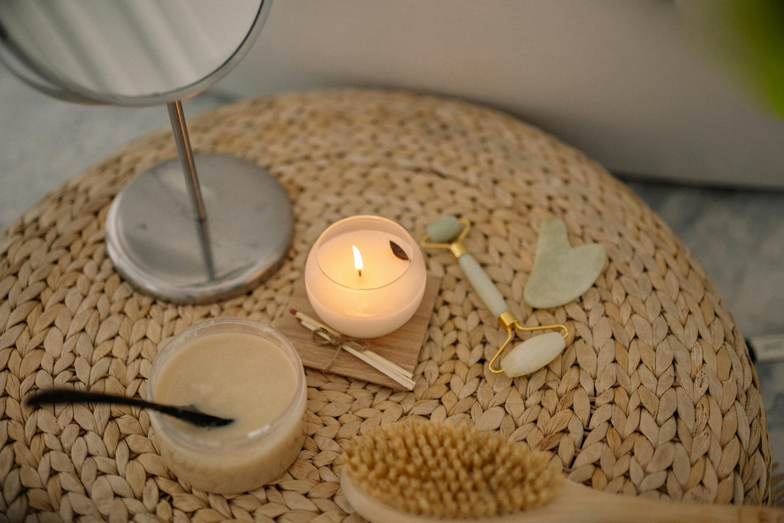 A serene spa setup featuring a lit candle, jade face roller, and body scrub on a woven mat.