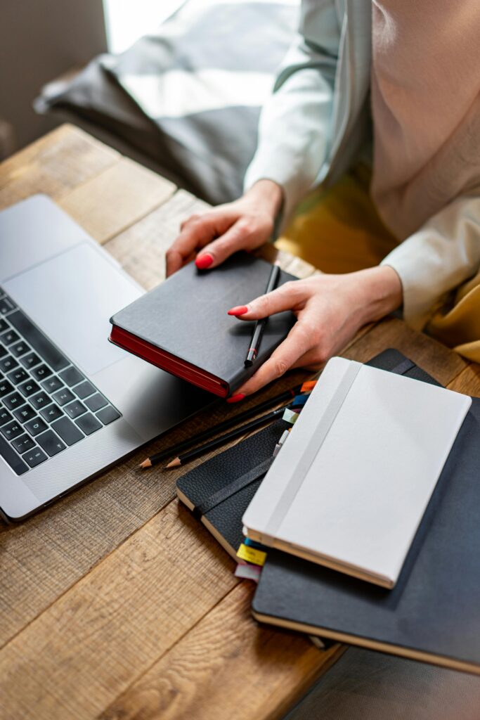 A woman organizing notebooks and journals at a wooden desk with a laptop.