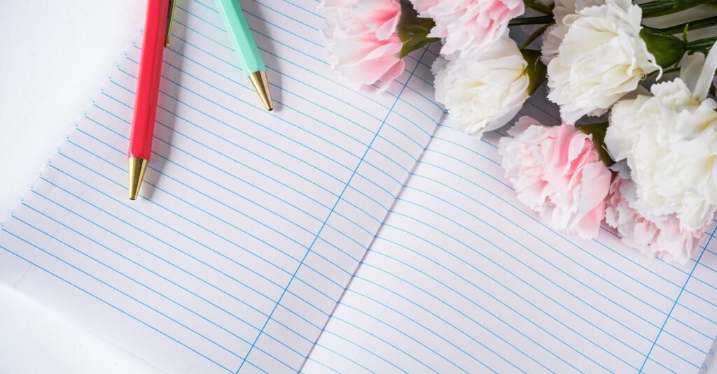 A flat lay featuring an open lined notebook with colorful pens and carnations, offering creative inspiration.