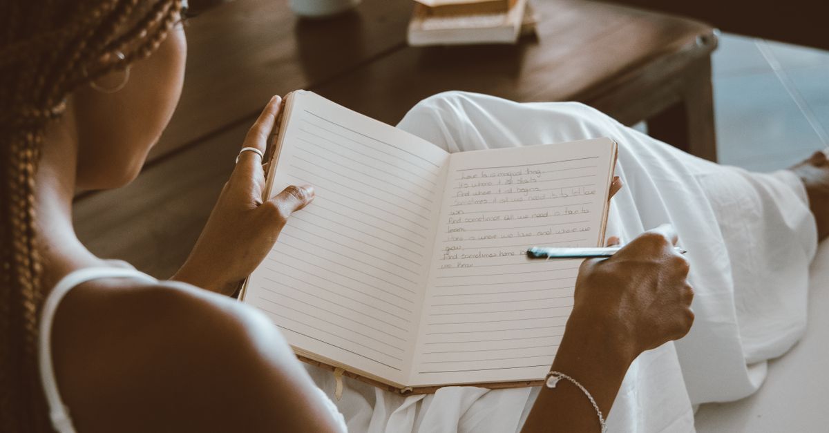 A black woman writes in a notebook, enjoying a calm indoor setting with natural light.