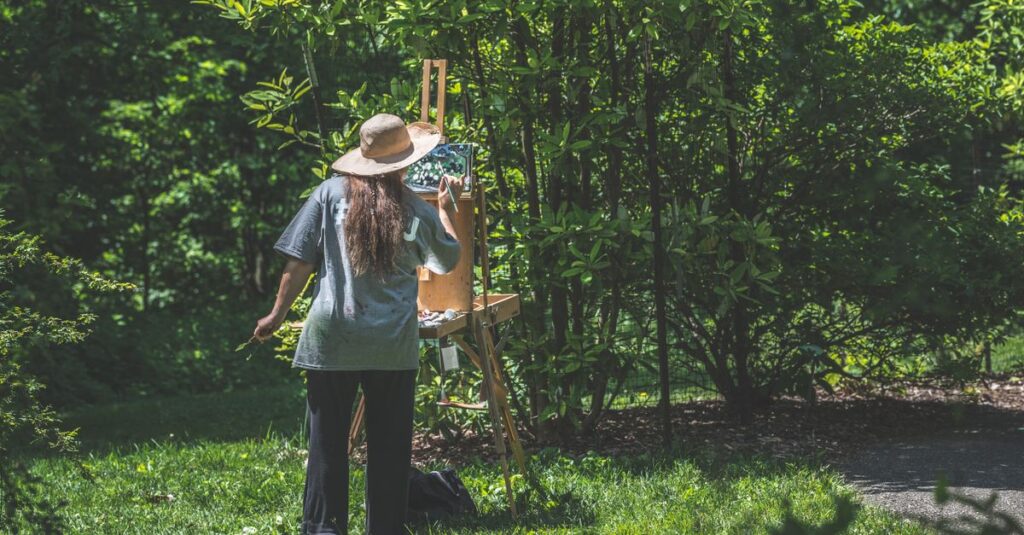 A woman with long hair paints on an easel in a lush, sunlit park.
