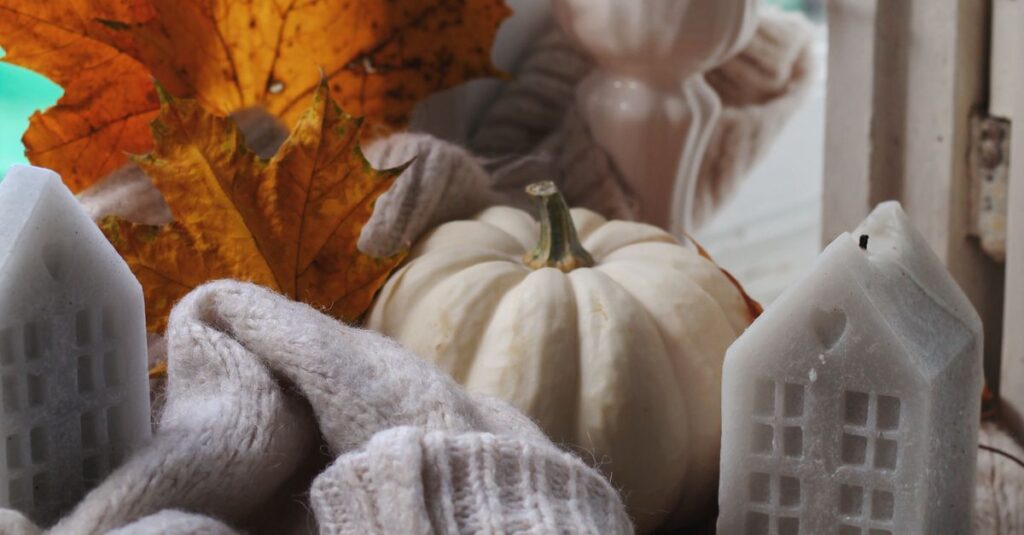 Warm and cozy autumn setting featuring a white pumpkin, candles, and fall leaves by a window.
