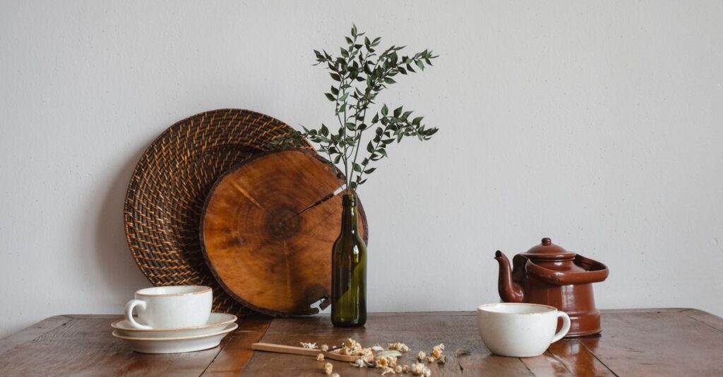 A rustic still life showcasing a teapot, cups, and a bottle with branches on a wooden table.