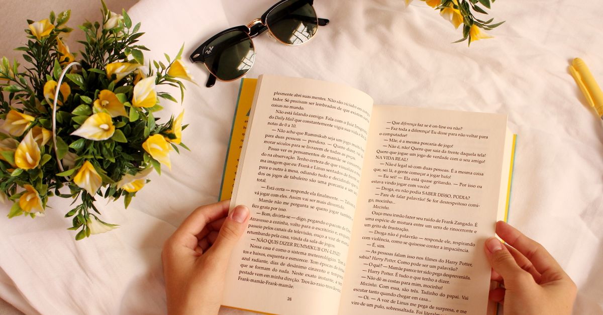 Hands holding a book surrounded by yellow flowers and sunglasses on a sunny day.