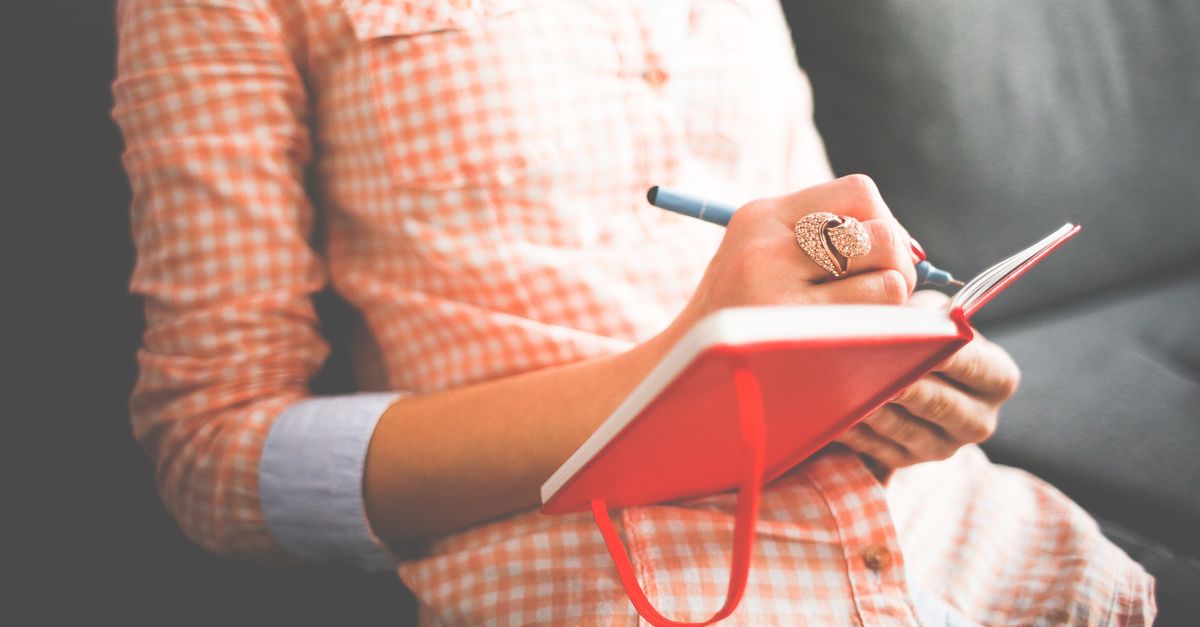 Close-up of a woman writing in a red journal on a sofa, wearing a stylish ring.
