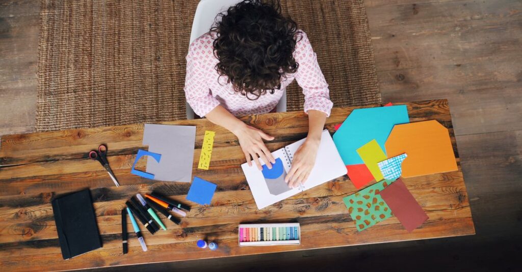 Person engaging in artistic crafts with colorful paper and tools on a wooden table.