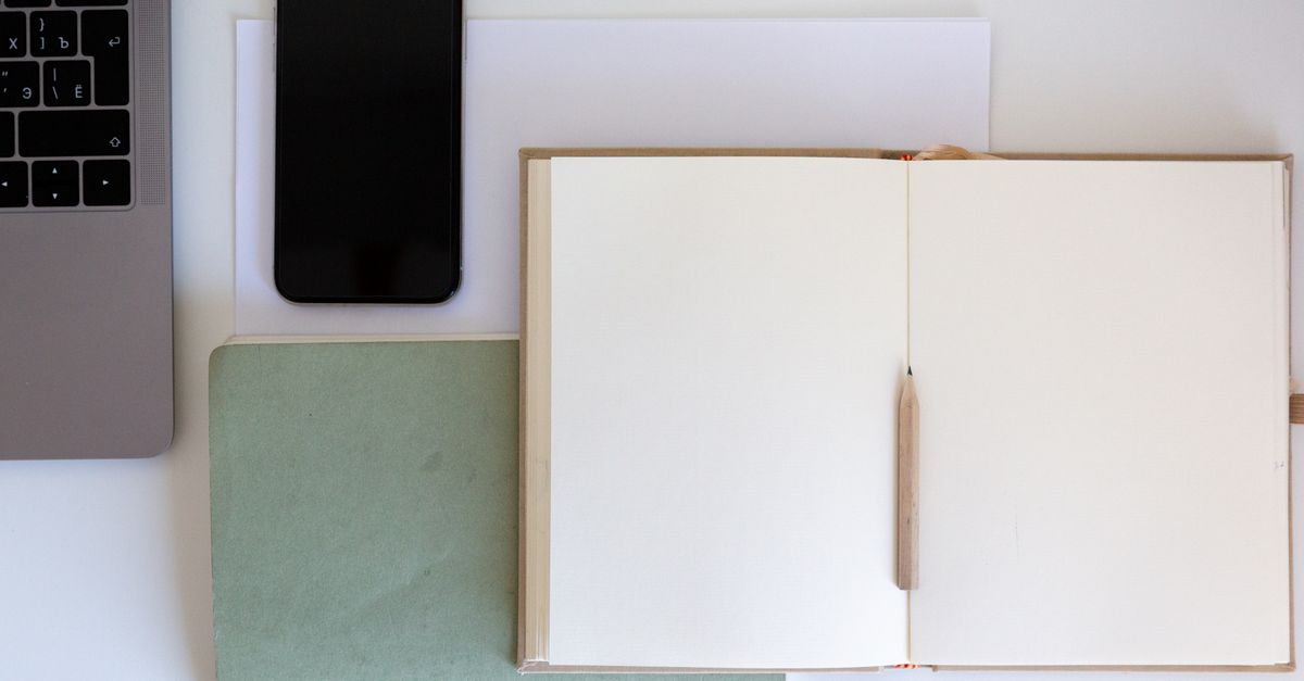 Top view of opened notebook near white paper and pencil placed on book near smartphone and laptop keyboard in office