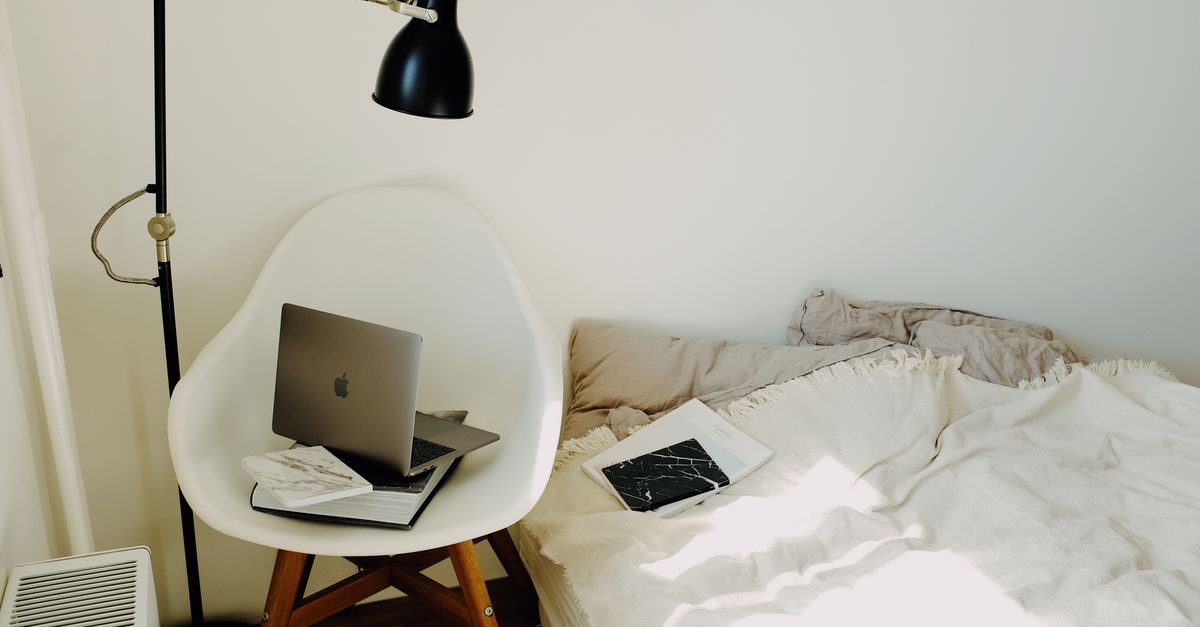 High angle of laptop placed on big black book on white armchair under lamp near comfortable bed in modern light bedroom at home