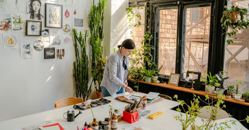 Side view of female designer creating drawings at desk with collection of felt pens and papers near wall with artworks and plants on windowsill in daylight
