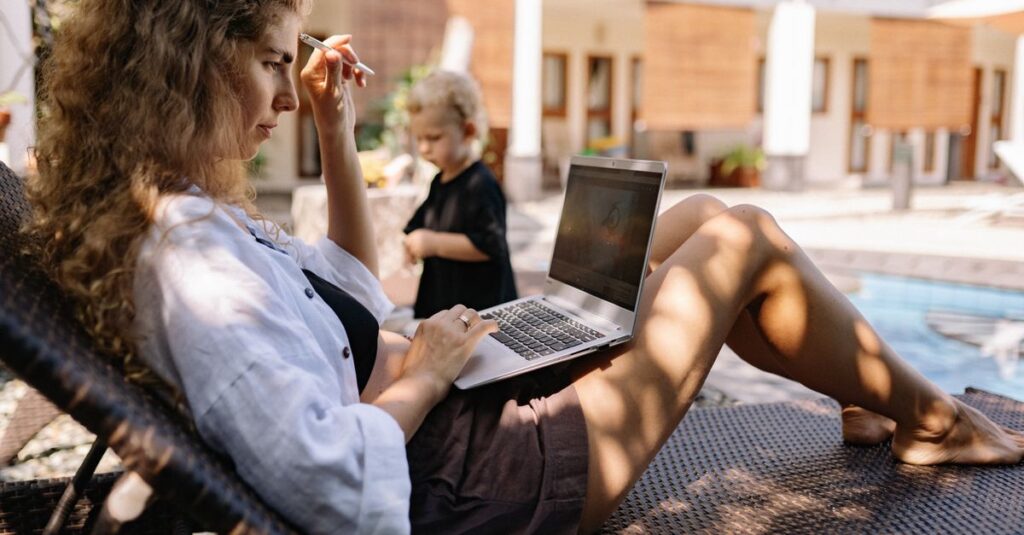 Woman typing on a laptop while relaxing outdoors on a sunlounger by the pool.