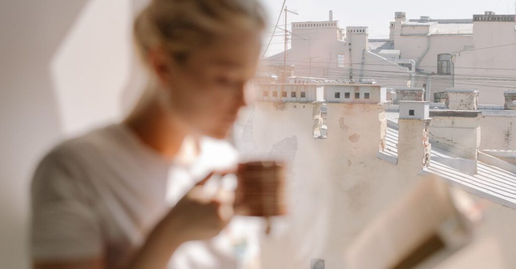 A woman sits and reads by a window, with a view of rooftops and clear skies.