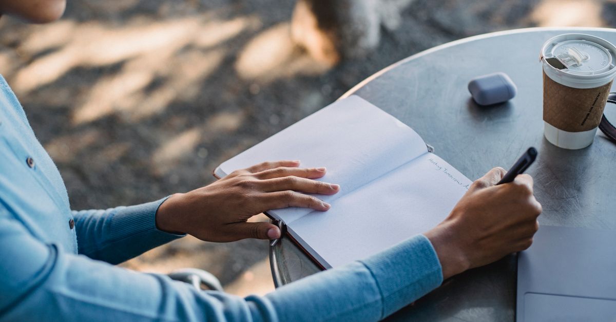 A close-up of a person writing notes in a notebook at an outdoor cafe table with a coffee cup.