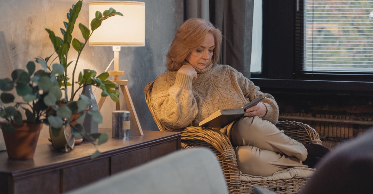 Senior woman in a sweater reading in a cozy room with plants and a lamp.