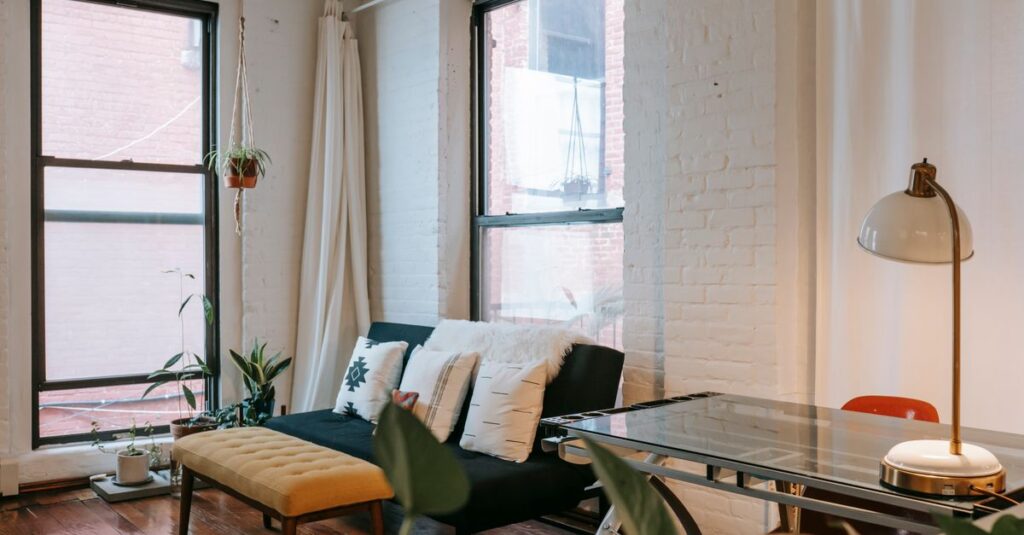 Soft blue couch with yellow pouf placed near windows in modern room with lamp on glass table and green plants