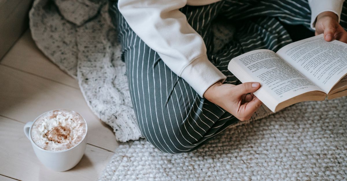 High angle of crop anonymous woman sitting on soft blanket on floor and reading literature with mug of coffee
