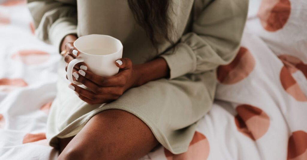 Crop anonymous ethnic female with cup of aqua sitting with crossed legs on bed blanket at home