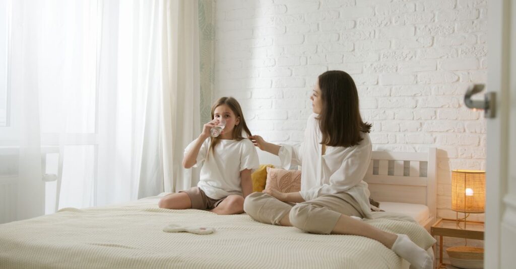 A mother and daughter share a warm moment sitting on a bed in a bright, cozy bedroom.