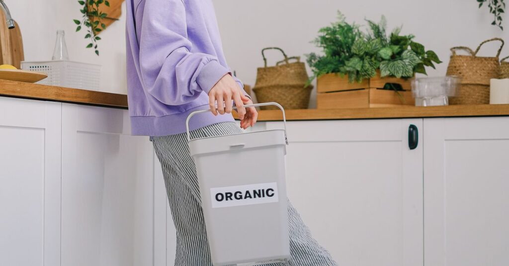 Side view of positive young female in trendy clothes standing with container with organic garbage in light room in daytime