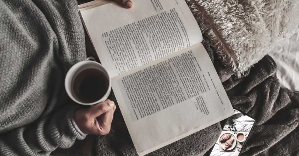 From above of crop anonymous female with mug of hot tea reading book under plaid near smartphone at home